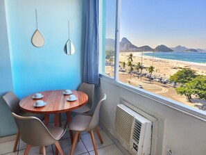 A room with a wooden table and chairs, a window with a view of a beach and mountains, and a wall-mounted air conditioner. - Ocean View Copacabana Best Spot (Rio de Janeiro)