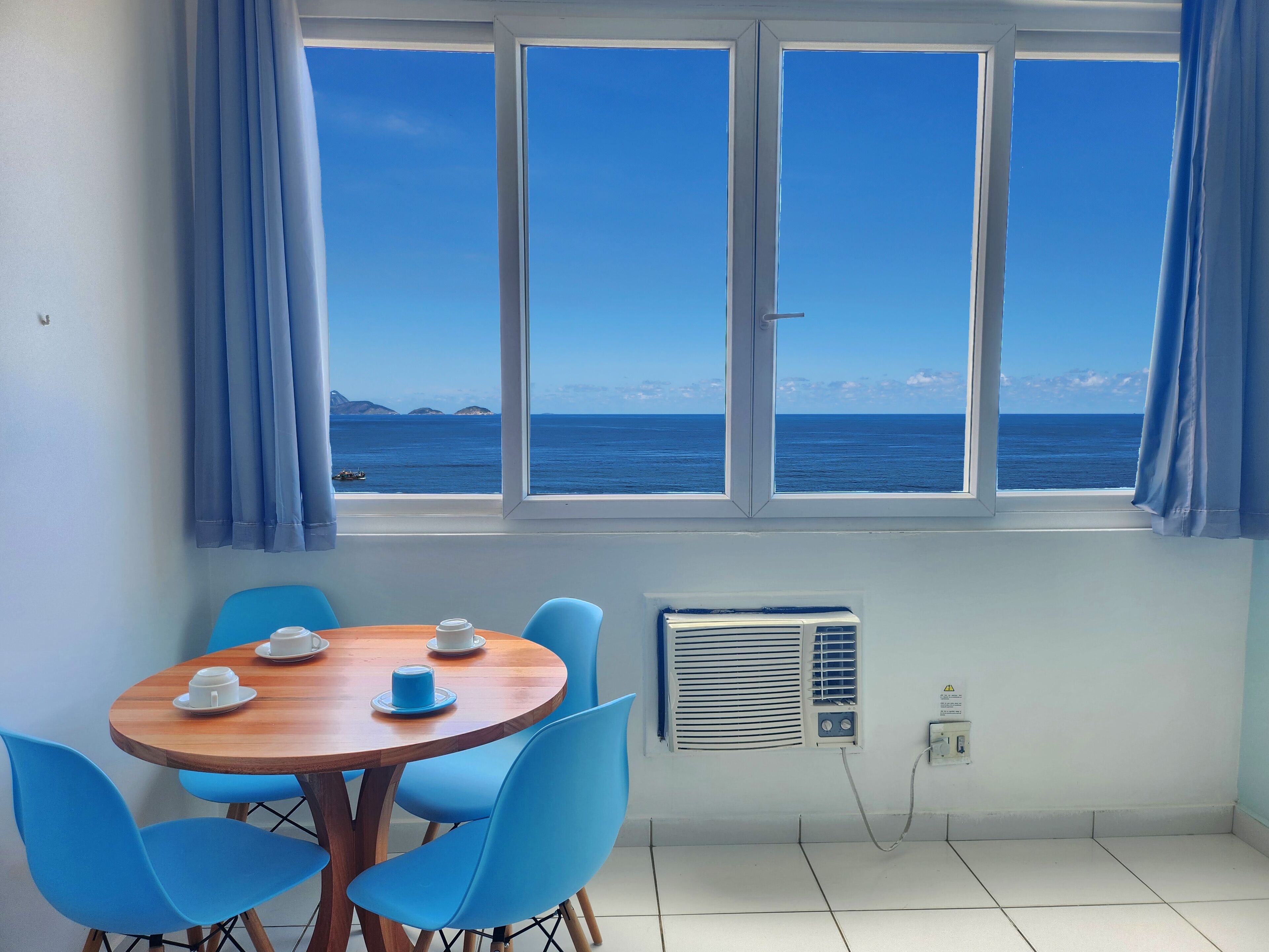 A room with a wooden table and blue chairs, a window with a view of the sea, and a wall-mounted air conditioner.