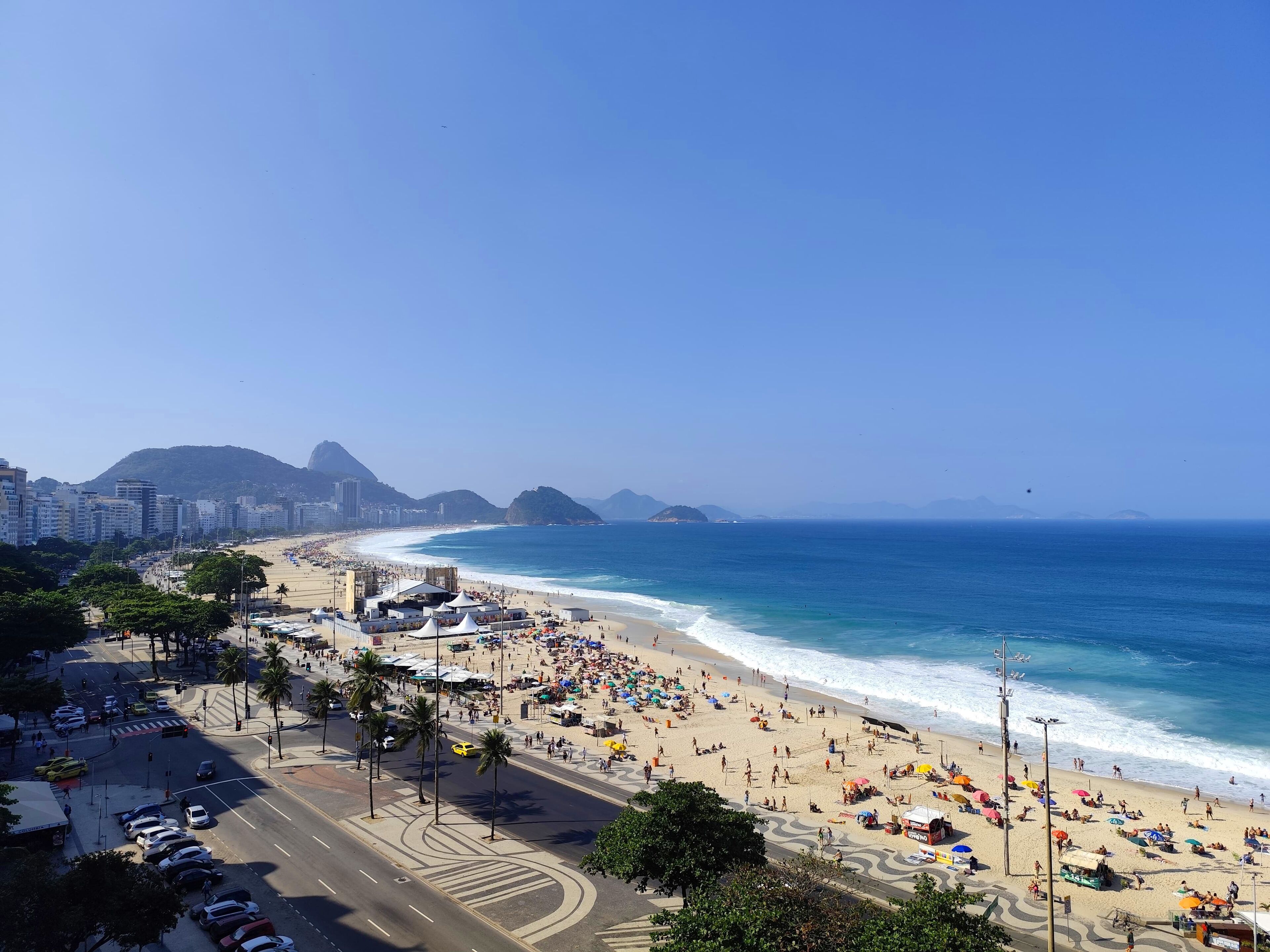 A beach with a road in the foreground, a cityscape in the background, and a clear sky.