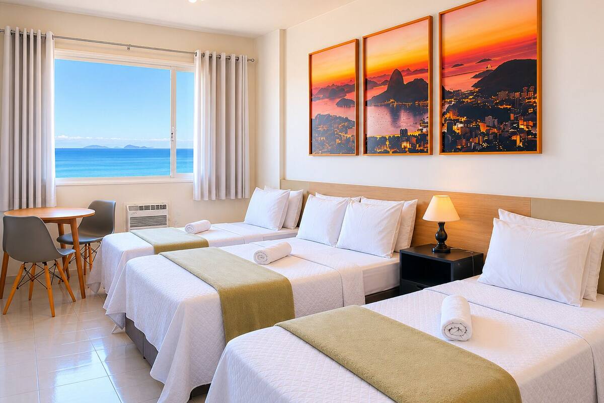 A hotel room with two beds, a desk, chairs, a view of the sea, and three sunset paintings on the wall.
