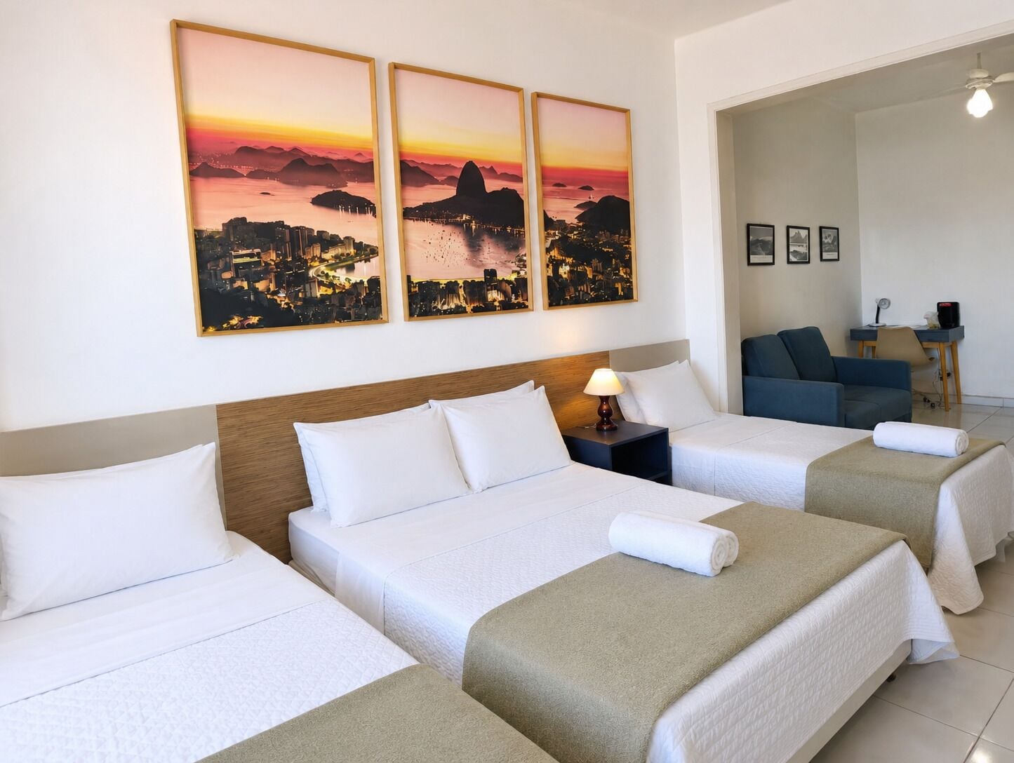 A hotel room with three beds, a desk, a chair, and a wall with three sunset cityscape paintings.