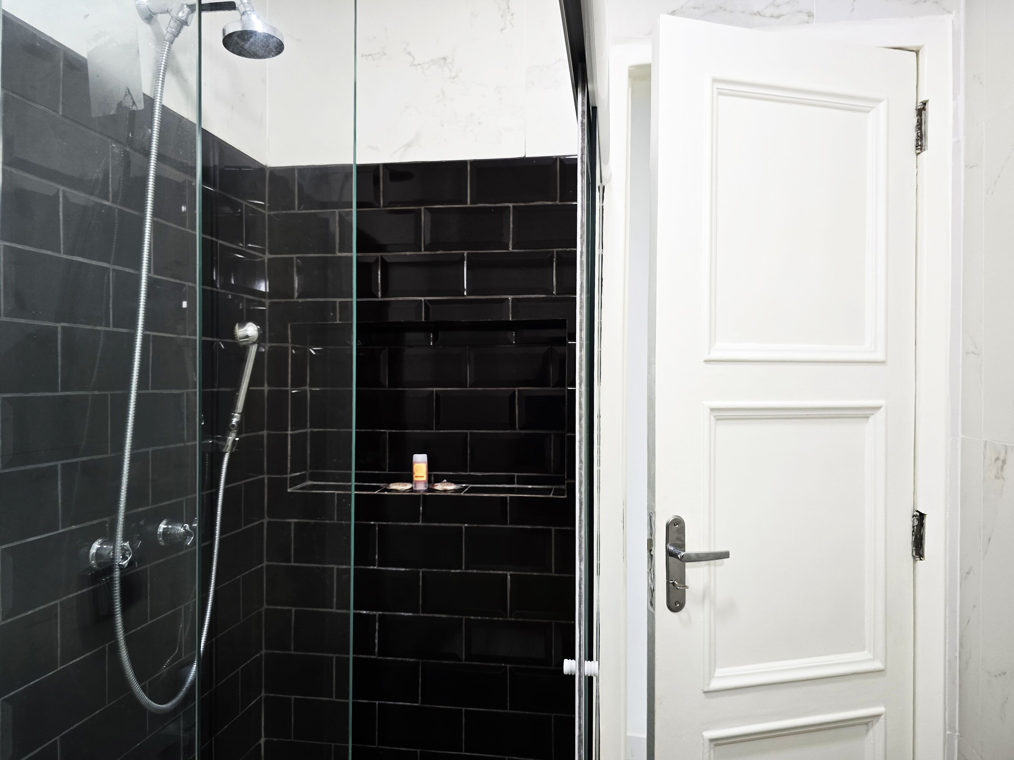 A bathroom with black tiled walls, a glass shower enclosure, a white door, and a small shelf with toiletries.
