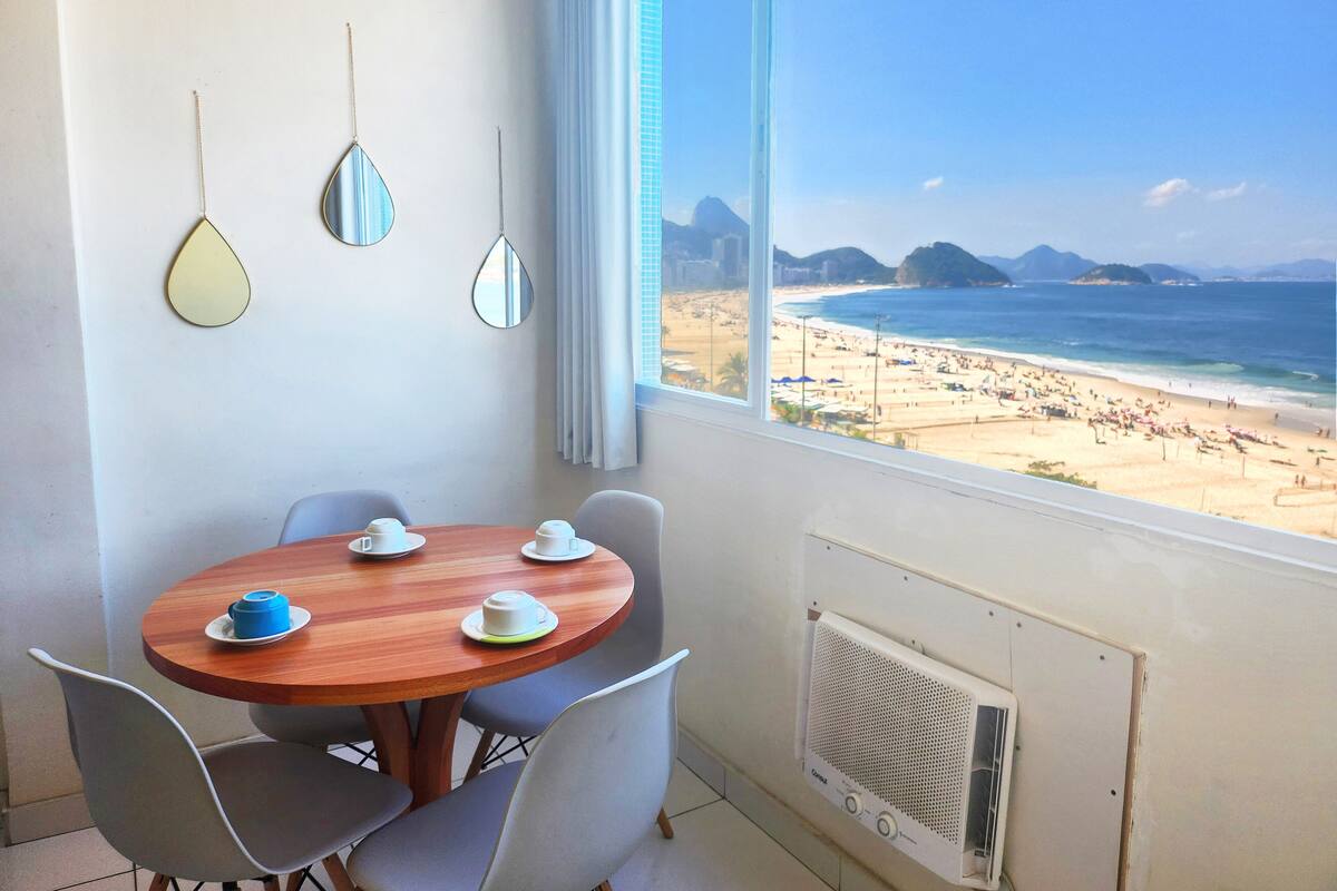A room with a wooden table, four chairs, a window with a beach view, and a wall-mounted air conditioning unit.