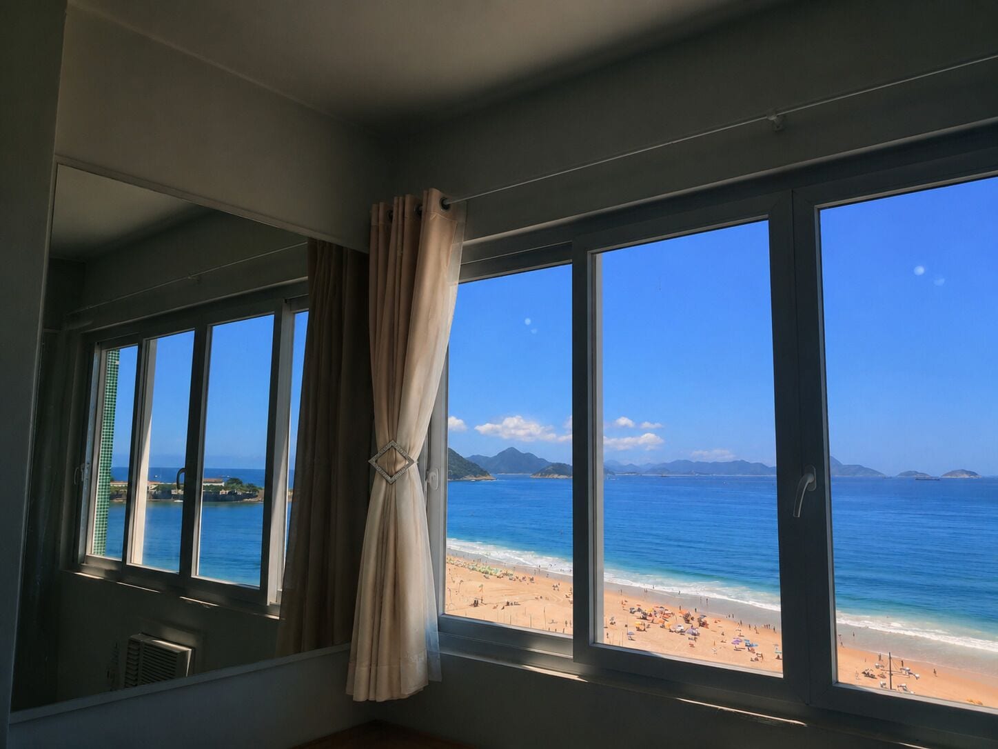 A room with large windows overlooking a beach, a curtain, and a view of the ocean and distant mountains.