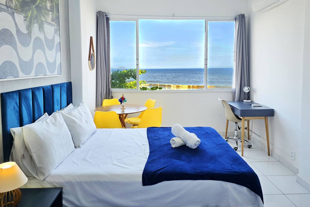 A hotel room with a bed, a desk, a chair, and a view of the sea.