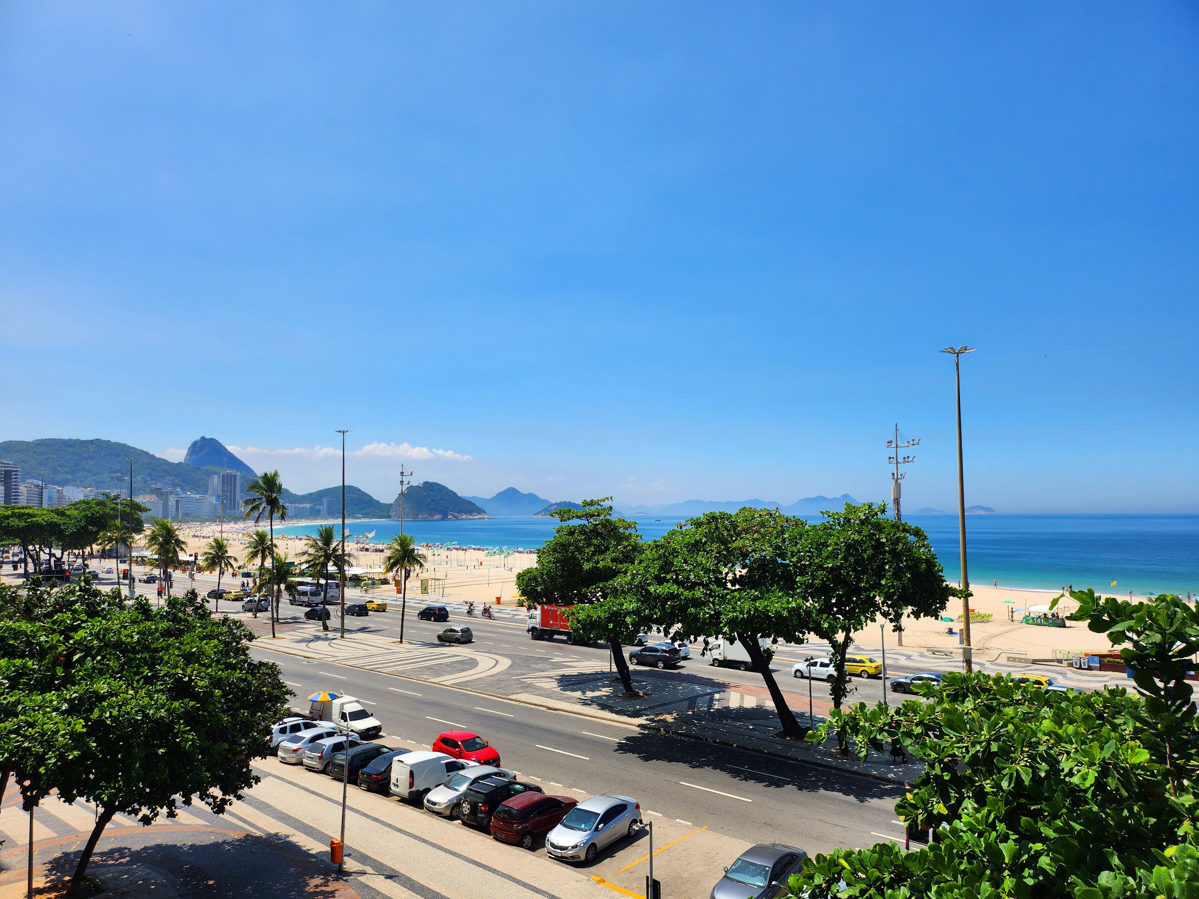 Un paysage urbain côtier avec une plage, des voitures garées le long de la rue et un ciel bleu dégagé.