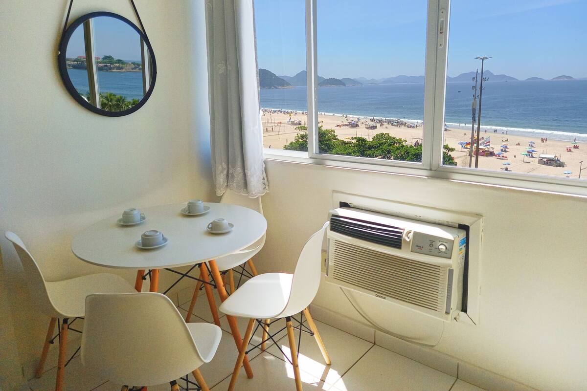 A room with a table and chairs, a window with a view of a beach, and a mirror on the wall.