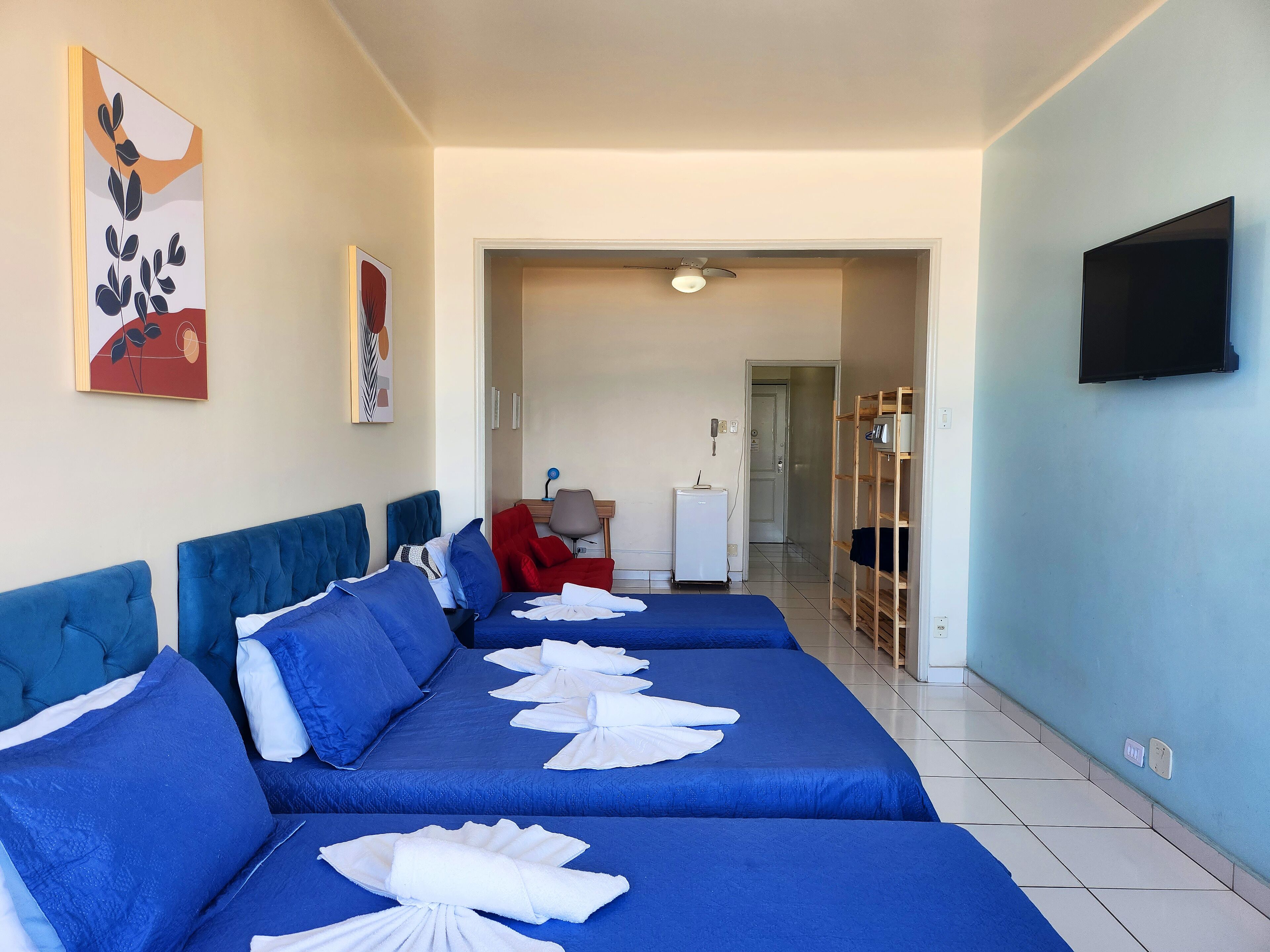 A hotel room with two blue beds, a TV, a desk with a chair, and a wardrobe.