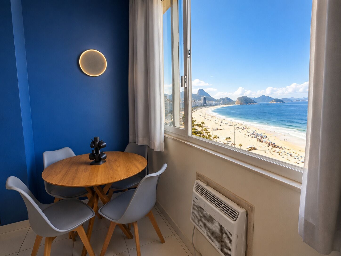 Una habitación con una mesa redonda de madera, dos sillas grises, un aire acondicionado de pared y una ventana con vista a la playa.