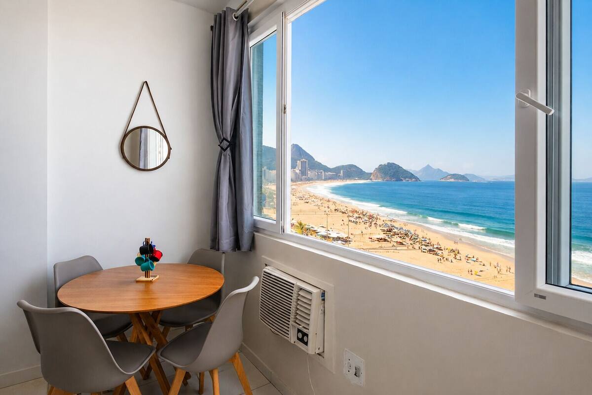 A room with a large window overlooking a beach, a round wooden table, and two chairs.
