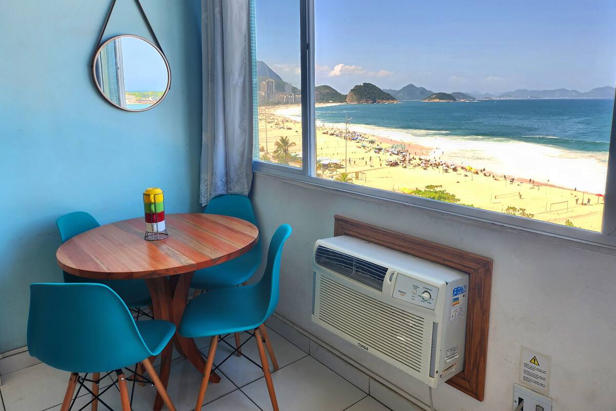 A room with a wooden table, blue chairs, a mirror, and a view of a beach with mountains.