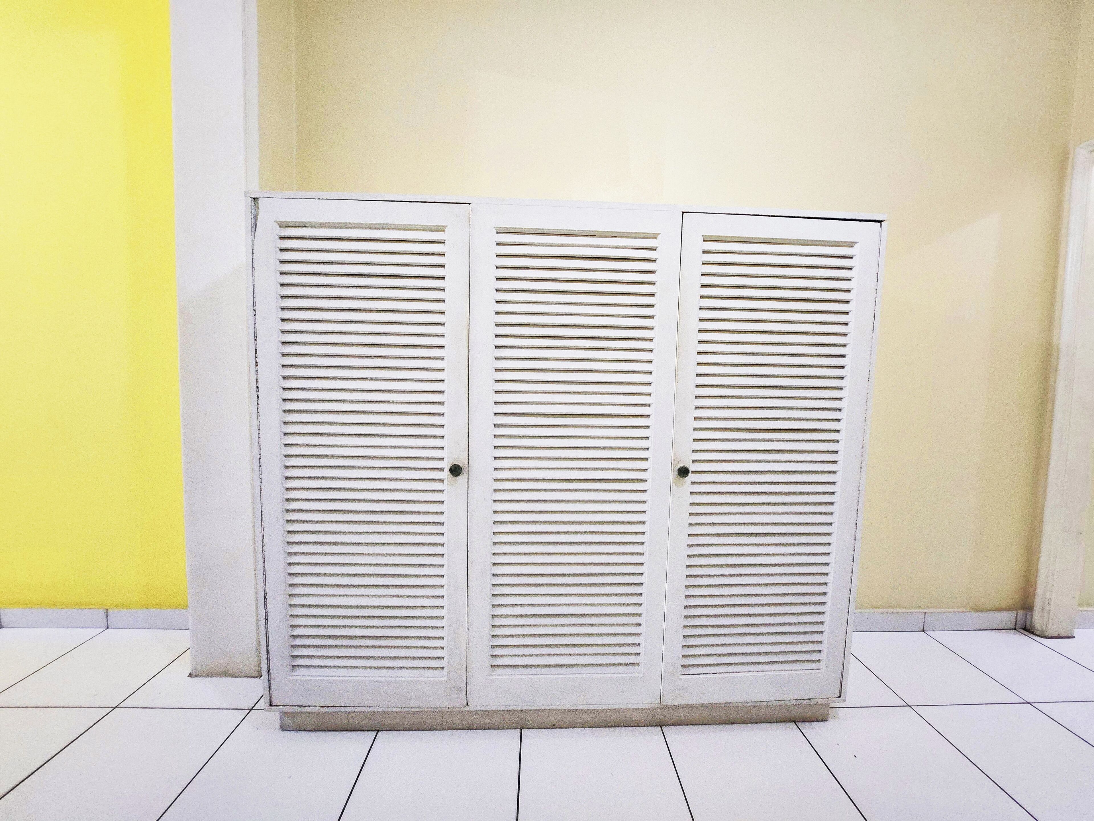A white wardrobe with horizontal slats, positioned against a yellow and beige wall, with a tiled floor.