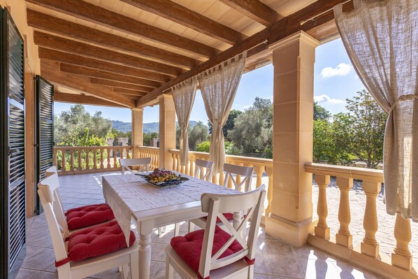 A covered outdoor dining area with a table set for two, white chairs with red cushions, and a view of greenery and hills in the background.