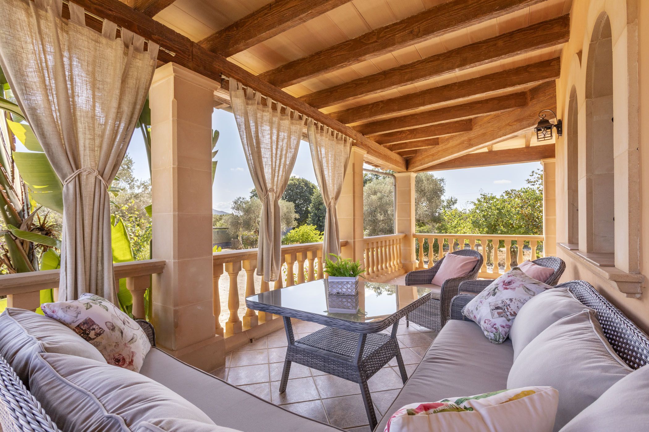 A covered outdoor seating area with wicker furniture, a glass table, and a view of greenery.