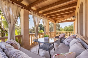 A covered outdoor seating area with wicker furniture, a glass table, and a view of greenery.