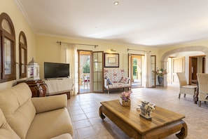 A spacious living room with a sofa, a wooden coffee table, and a television. There are two chairs and a patterned armchair. The room has a tiled floor and large windows with curtains. - Quiet villa with 4 bedrooms, 2 bathrooms, barbecue, wifi and pool (Inca)
