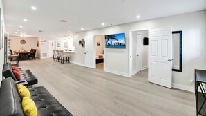 Een ruime woonkamer met een eettafel, een bank met kussens en een aan de muur bevestigde televisie.