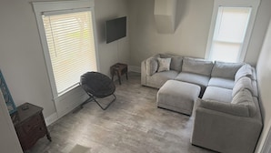 A living room with a sectional sofa, a black chair, a small wooden table, and a TV mounted on the wall. - Simba’s House Mid 1800’s Burton Village Retreat (Burton)