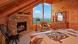 A cozy log cabin bedroom with a stone fireplace, a large window offering a scenic view, a quilted bedspread, and a wooden dresser. - Above It All #38 (Sevierville)