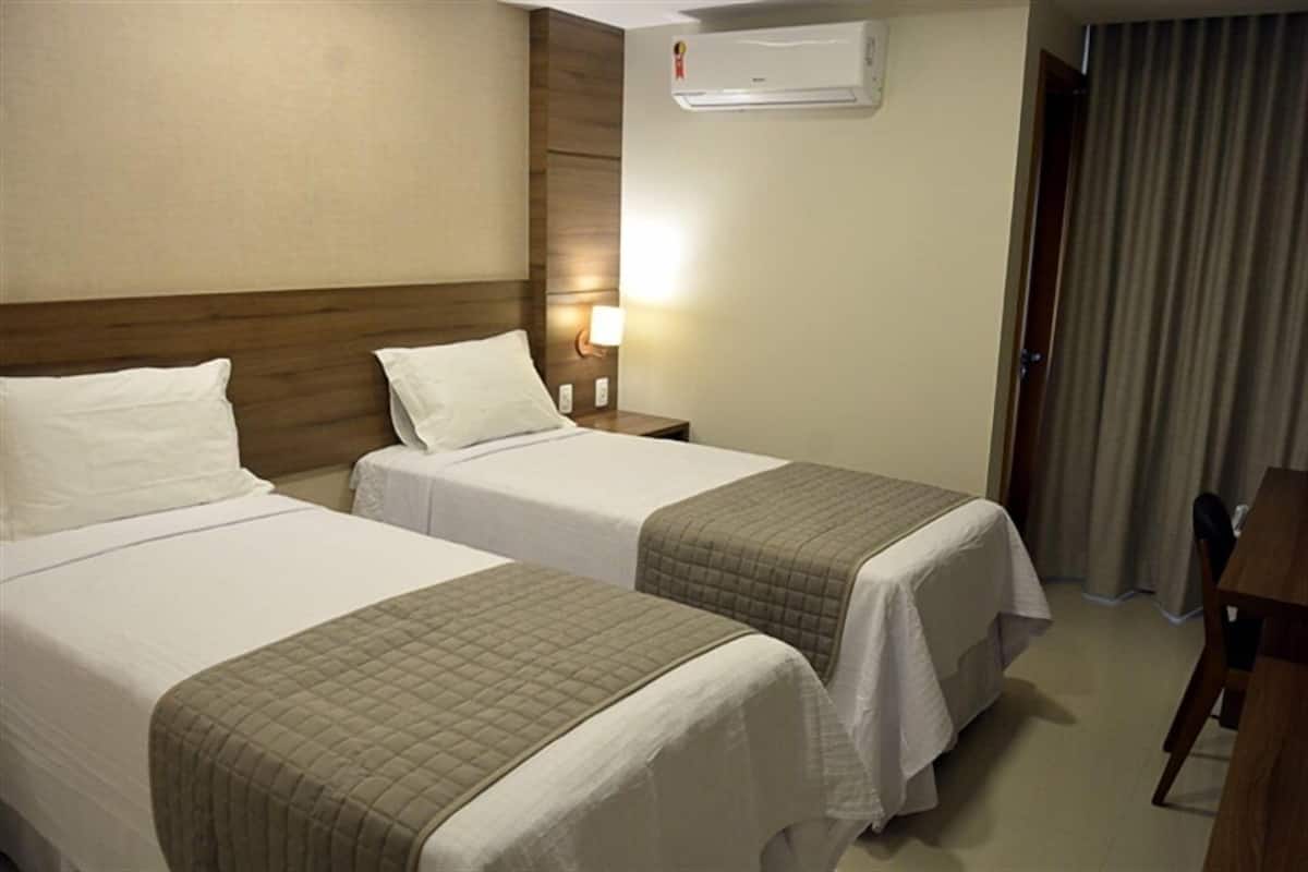 Habitación de hotel con dos camas, un escritorio, una silla, aire acondicionado y una mesita de noche con lámpara.