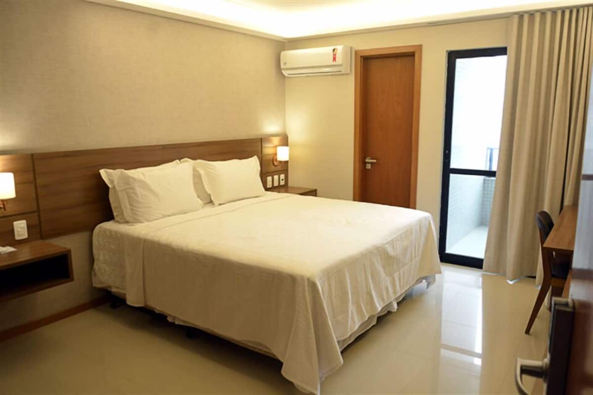 Habitación de hotel con una cama grande, una mesita de noche con lámpara, equipo de aire acondicionado, puerta a un balcón y vistas al exterior.