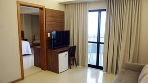A hotel room with a bed, a desk with a television, a small refrigerator, a sofa, and a balcony with curtains. - Pituba Praiamar Hotel (Salvador)