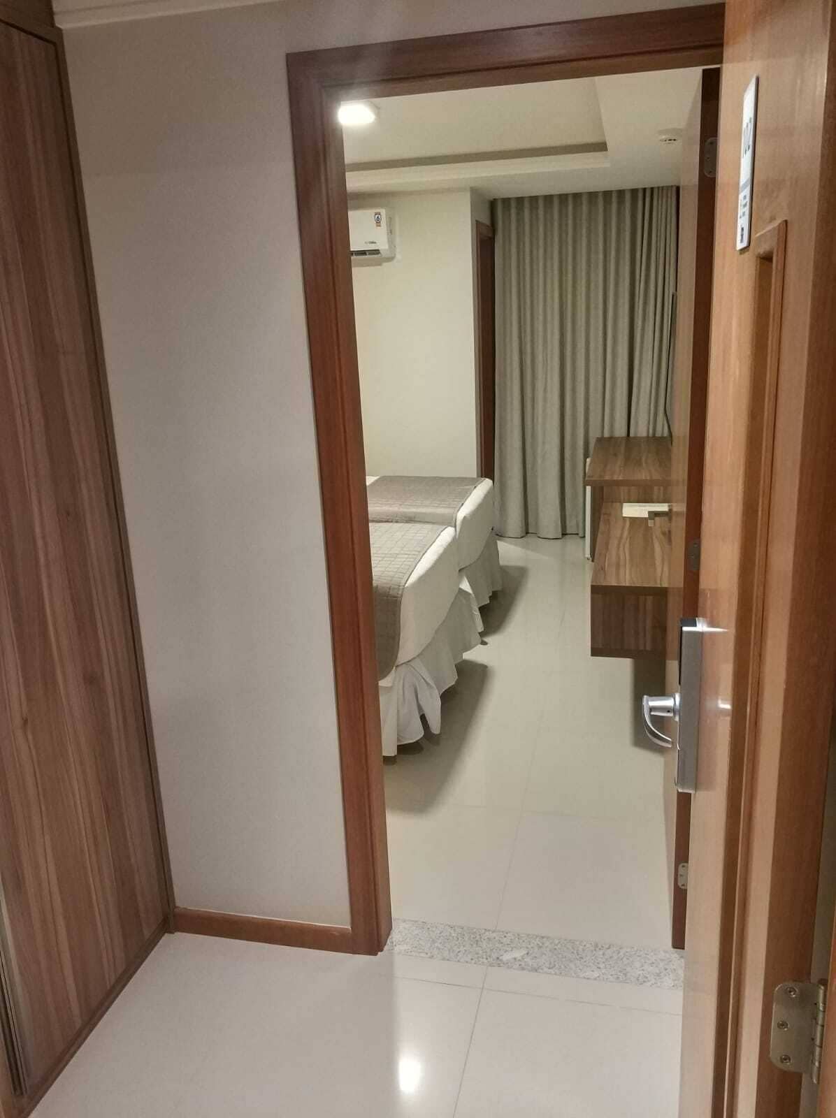 Une chambre d’hôtel avec une porte en bois, un lit avec une literie blanche, une petite table de chevet en bois et un climatiseur fixé au mur.