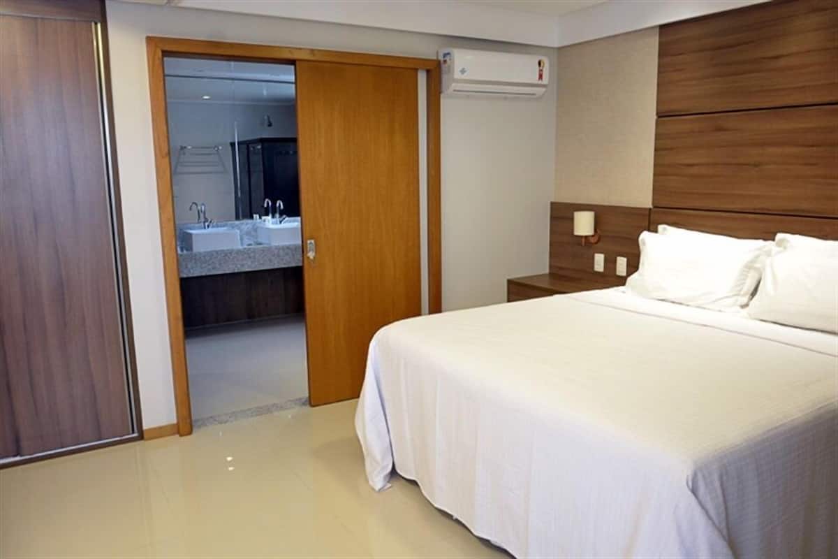 Habitación de hotel con cama, cabecera de madera, mesita de noche y baño visible a través de una puerta abierta.