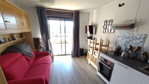 A compact living space with a red sofa, wooden cabinets, a kitchen area with a stove, and a sliding glass door leading to an outdoor area.