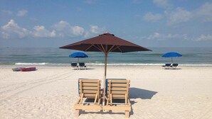 Una playa con dos sillones de madera bajo una sombrilla marrón, y dos sombrillas azules al fondo.