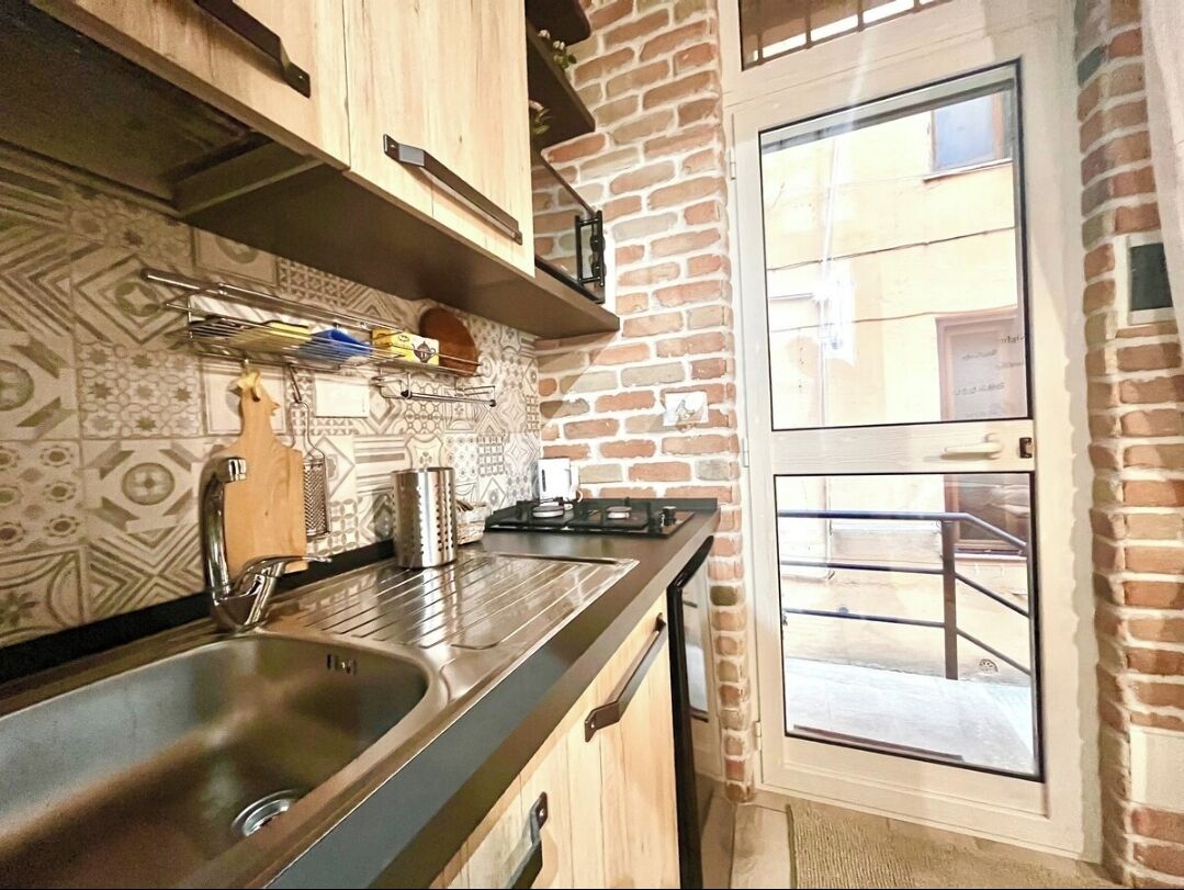 A kitchen with a stainless steel sink, wooden cabinets, a hanging rack, and a brick wall.