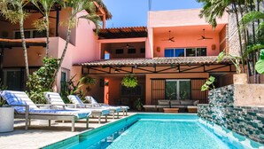 Una piscina con lettini, una zona relax coperta e un edificio con tetto in tegole e balconi.