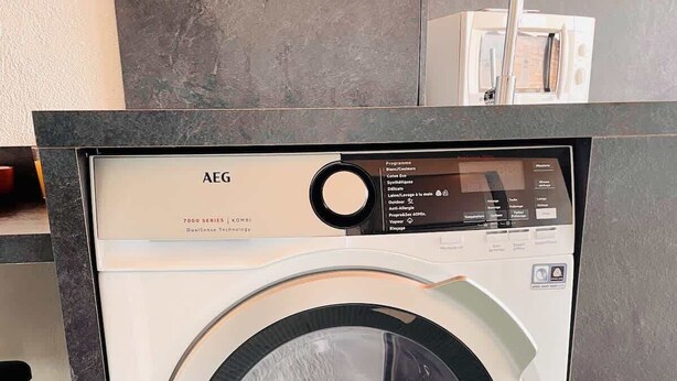 Una lavadora de carga frontal con un panel de control, ubicada en una cocina moderna con piso de baldosas y una encimera.