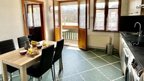 A dining area with a table set for breakfast, a kitchen with appliances, and a view through a door. - Angèle (Bains-les-Bains)