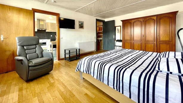 A bedroom with a bed, a reclining chair, wooden cabinets, and a kitchen area in the background. - Angèle (Bains-les-Bains)