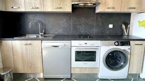 A modern kitchen with wooden cabinets, a dishwasher, an oven, and a washing machine. - Angèle (Bains-les-Bains)