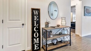 A hallway with a 'WELCOME' sign, a mirror, a console table with decorative items, and a bookshelf.