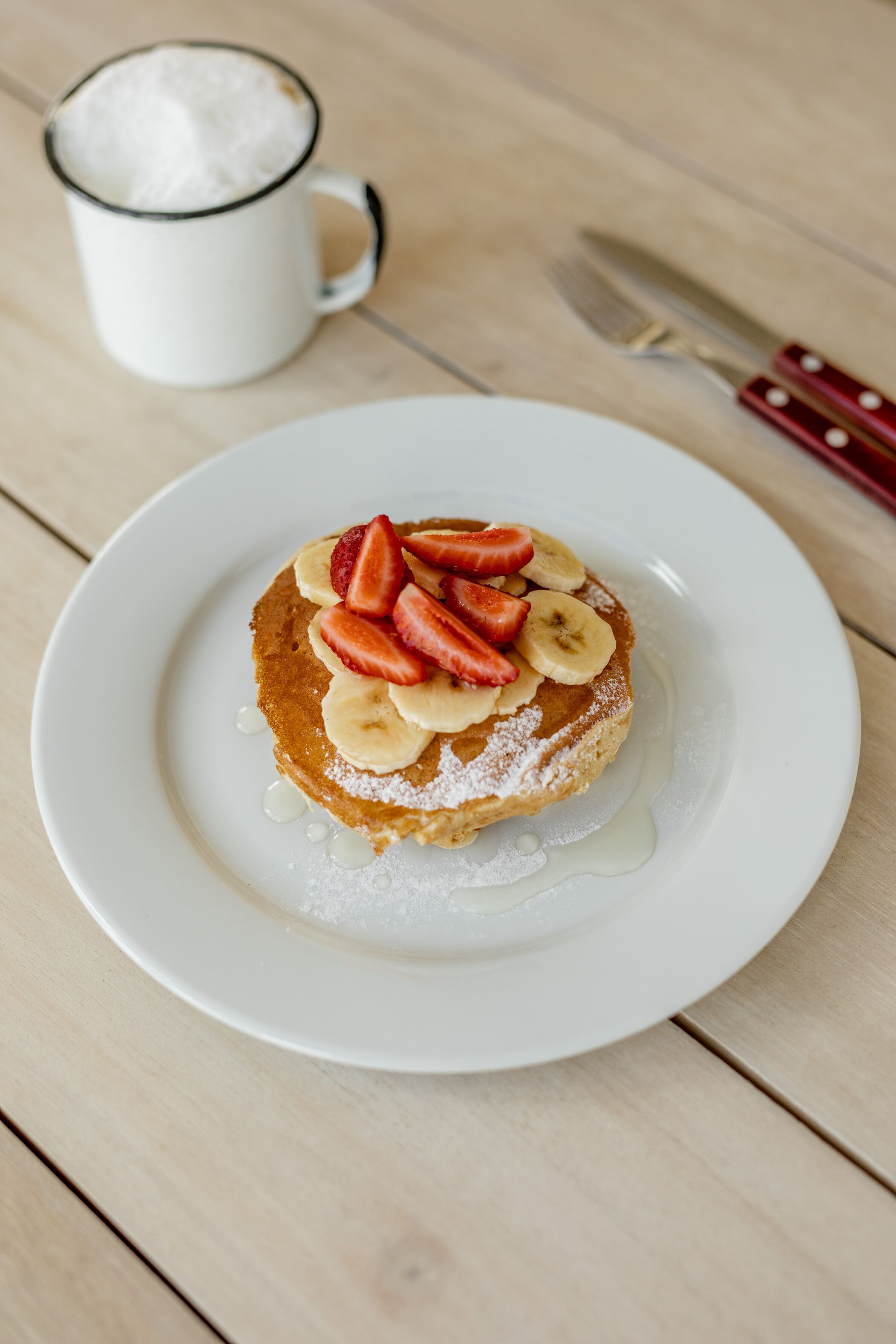 A plate with pancakes topped with strawberries and bananas, a white cup, and a knife on a wooden table.