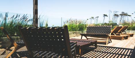A wooden deck with lounge chairs and a table, overlooking a grassy area with tall plants.