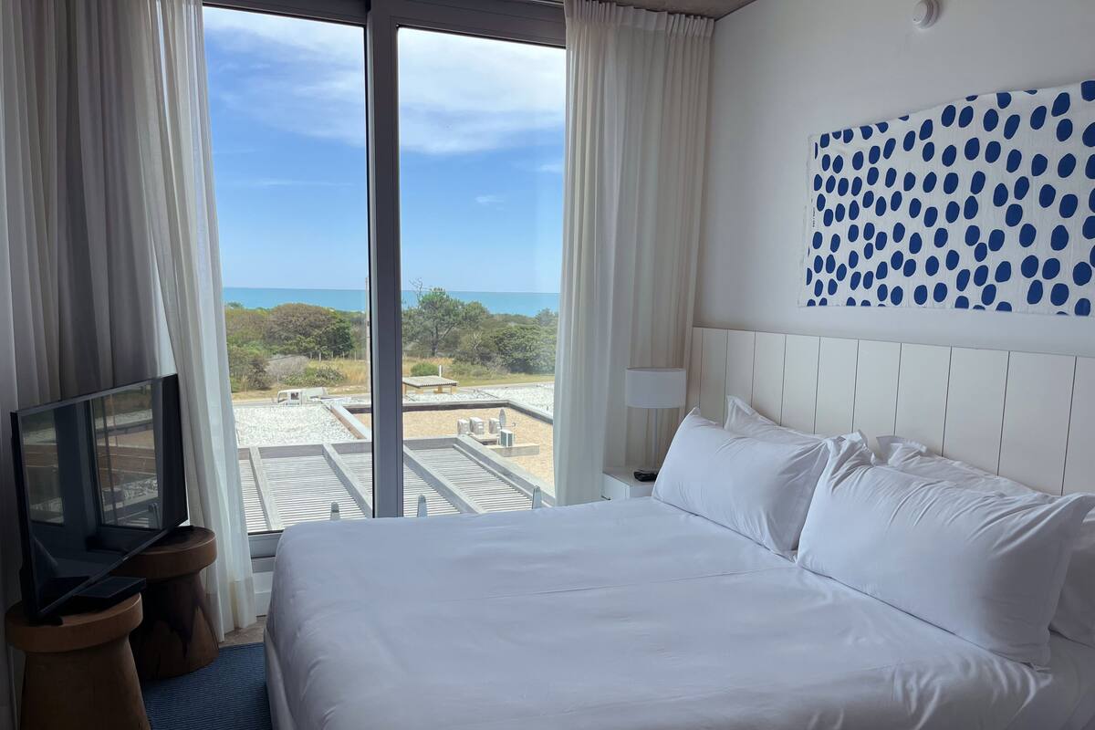 A hotel room with a large bed, a TV on a wooden side table, a view of the beach, and a polka dot wall decoration.