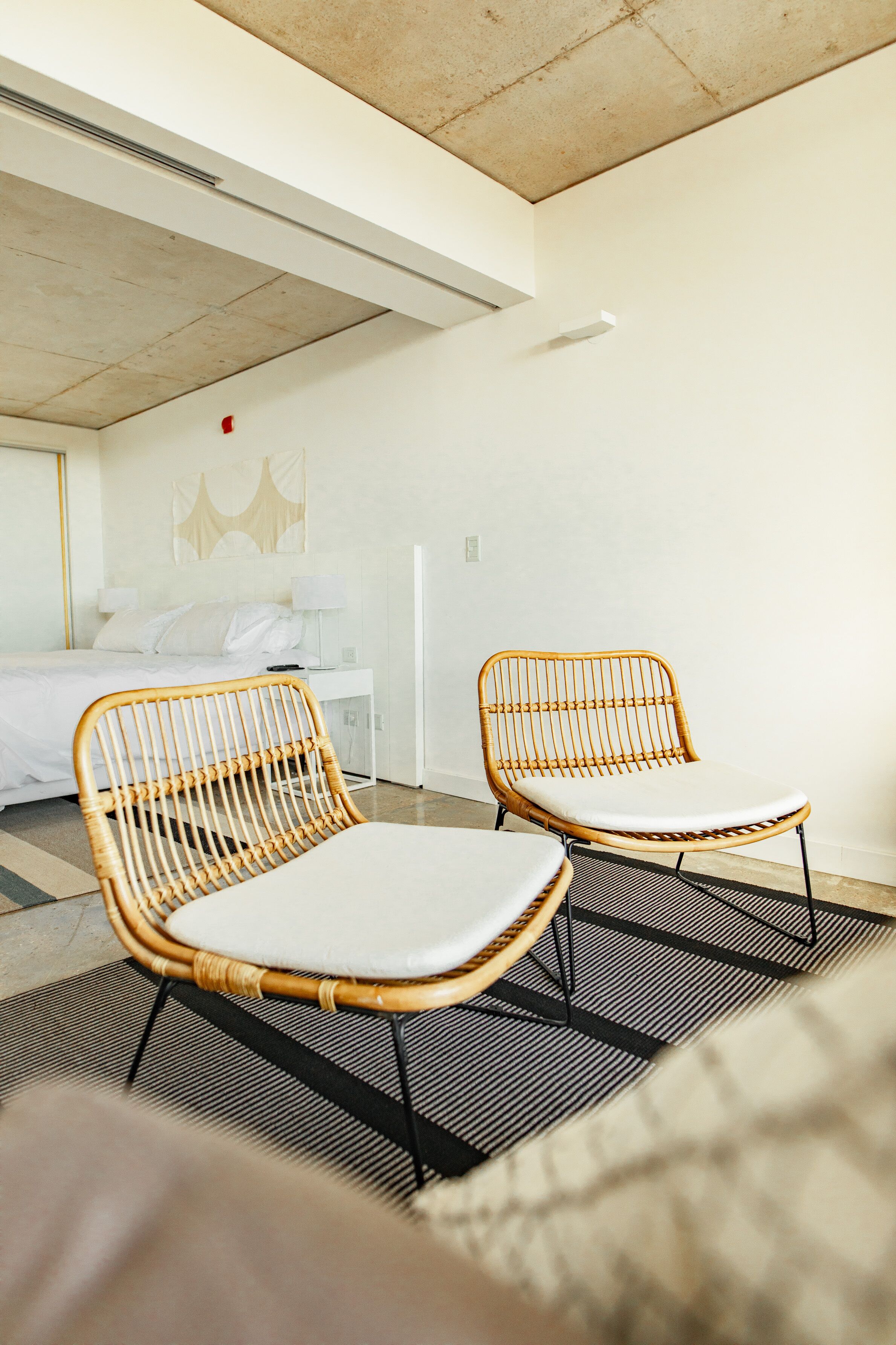 A modern hotel room with a bed, two wicker chairs, a white desk, and a large window.