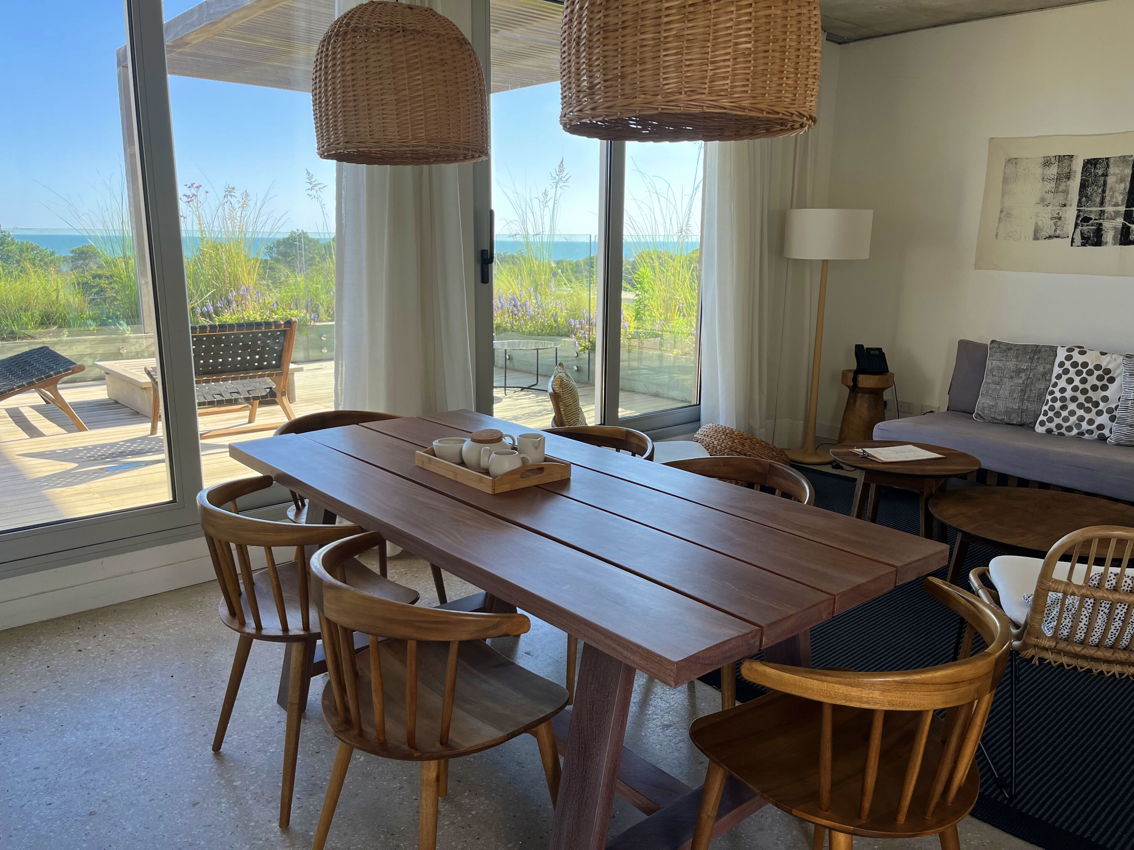 A dining area with a wooden table and chairs, a wicker hanging light fixture, and a view of the sea through large windows.
