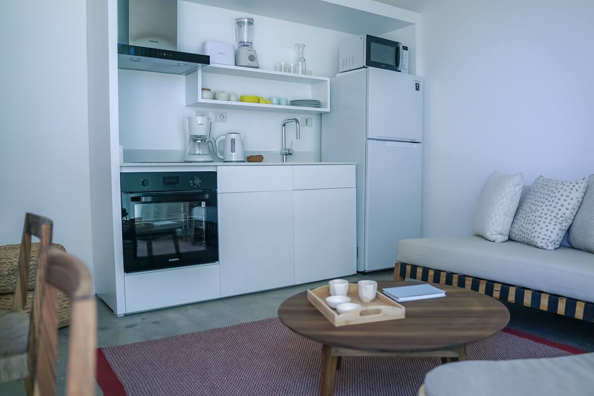 A compact kitchen with a microwave, oven, and coffee maker. A small dining table with cups and a notebook is in the foreground.
