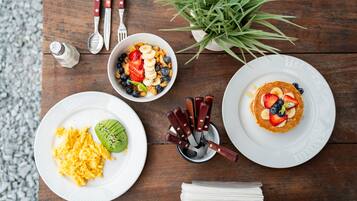 A wooden table with breakfast items including scrambled eggs, a green smoothie, and pancakes topped with fruits.