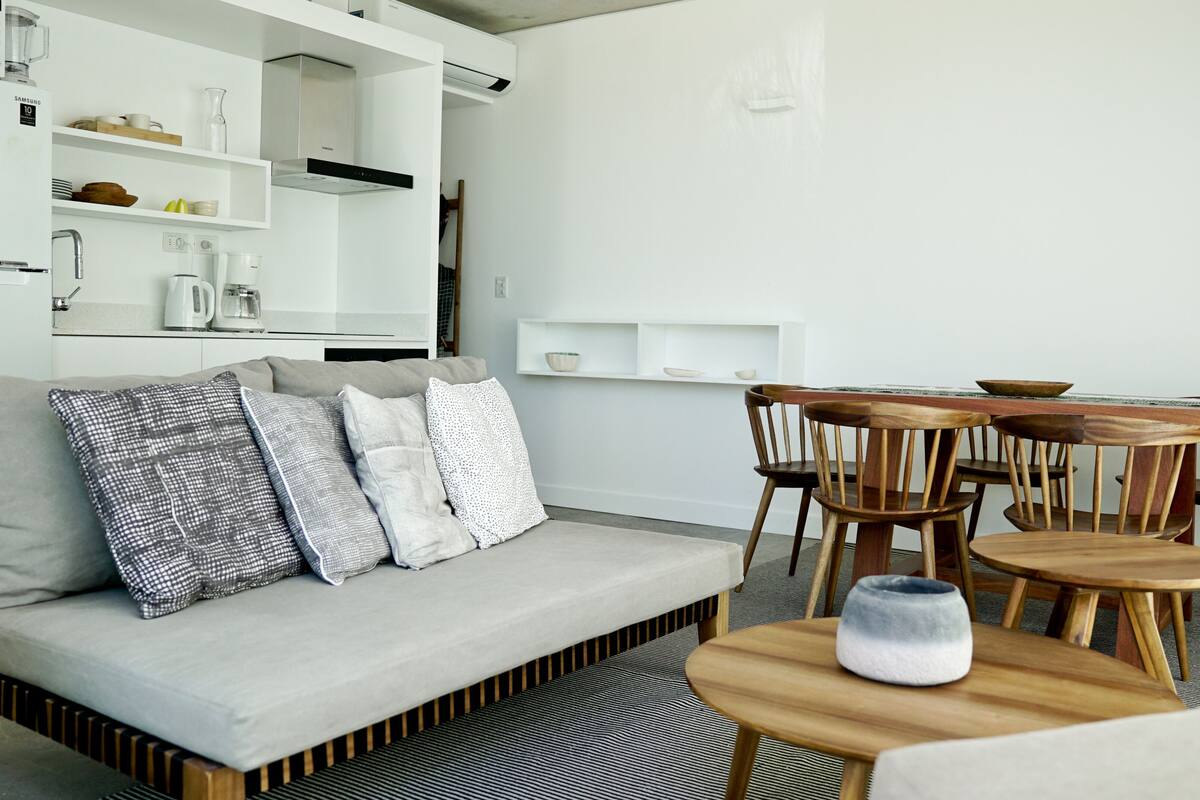 A modern living room with a sofa, wooden chairs, a coffee table, and a kitchen area with white cabinetry and shelves.