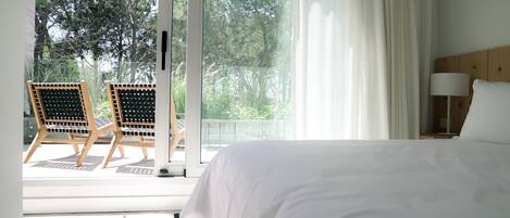 A hotel room with a large bed, a wooden chair, a small table, and a view of trees through a glass door.