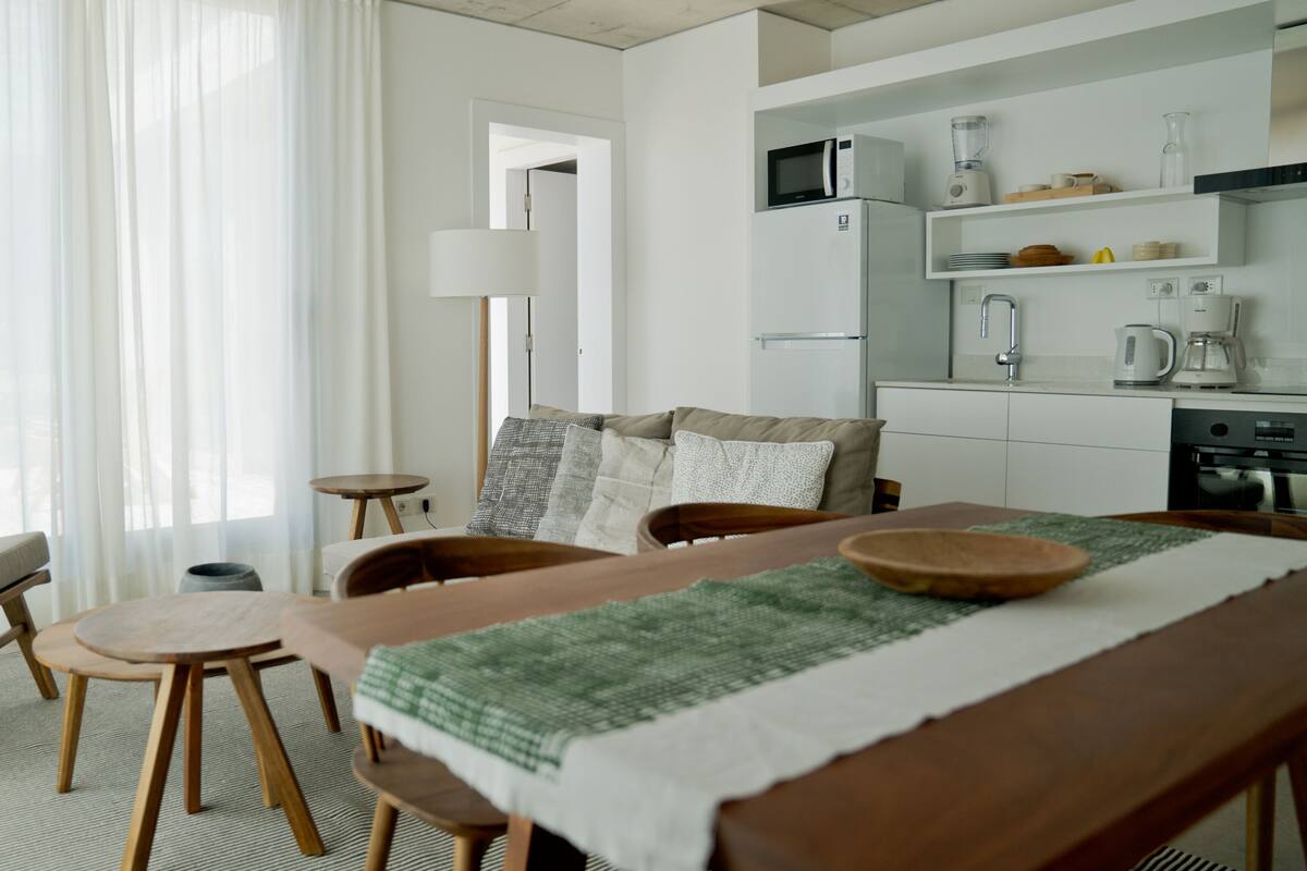 A modern kitchen with white appliances, a dining area with a wooden table and chairs, and a living area with a sofa and a lamp.