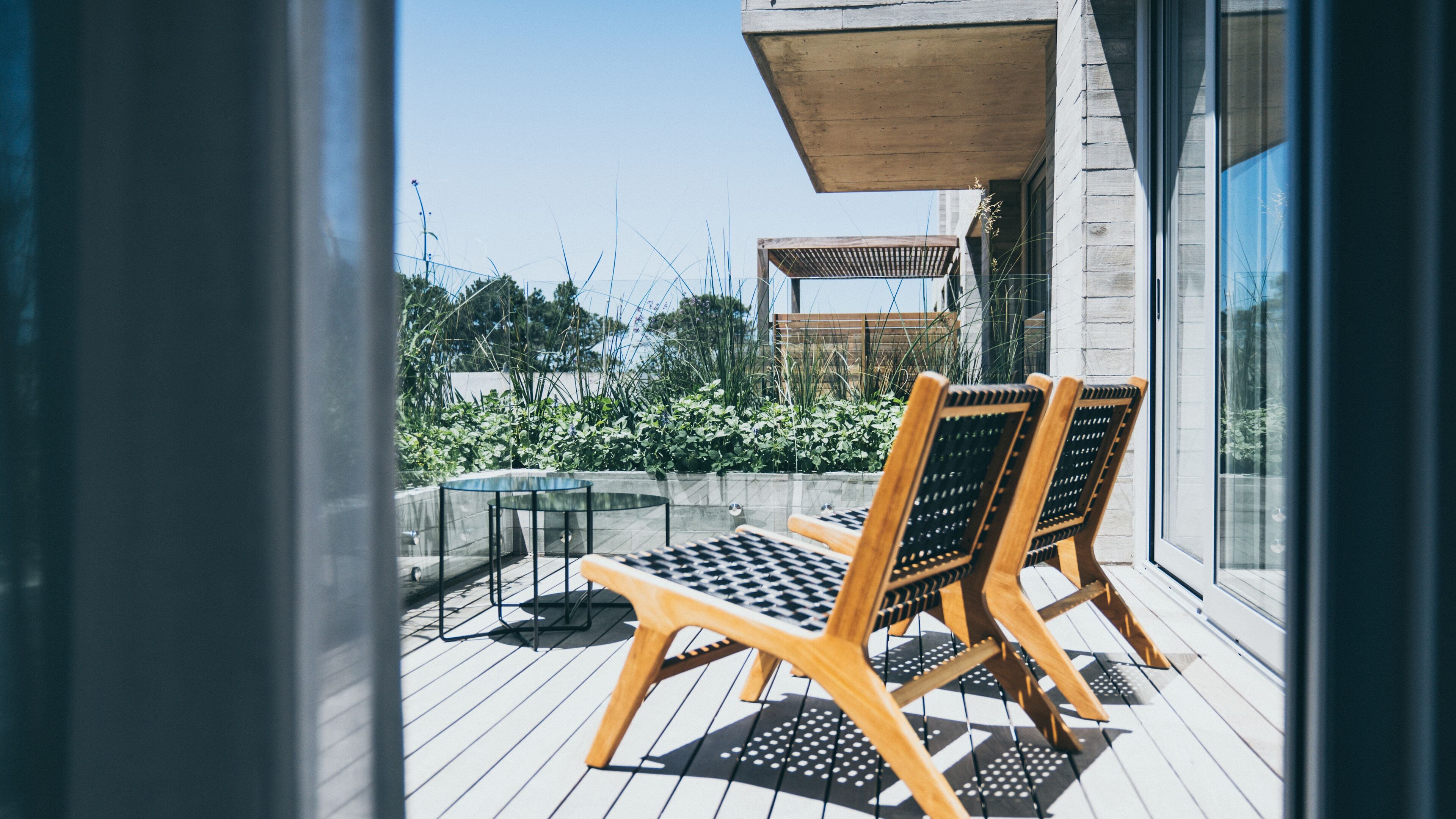 A wooden deck with two lounge chairs and a small table, overlooking a garden with various plants and a wooden pergola.