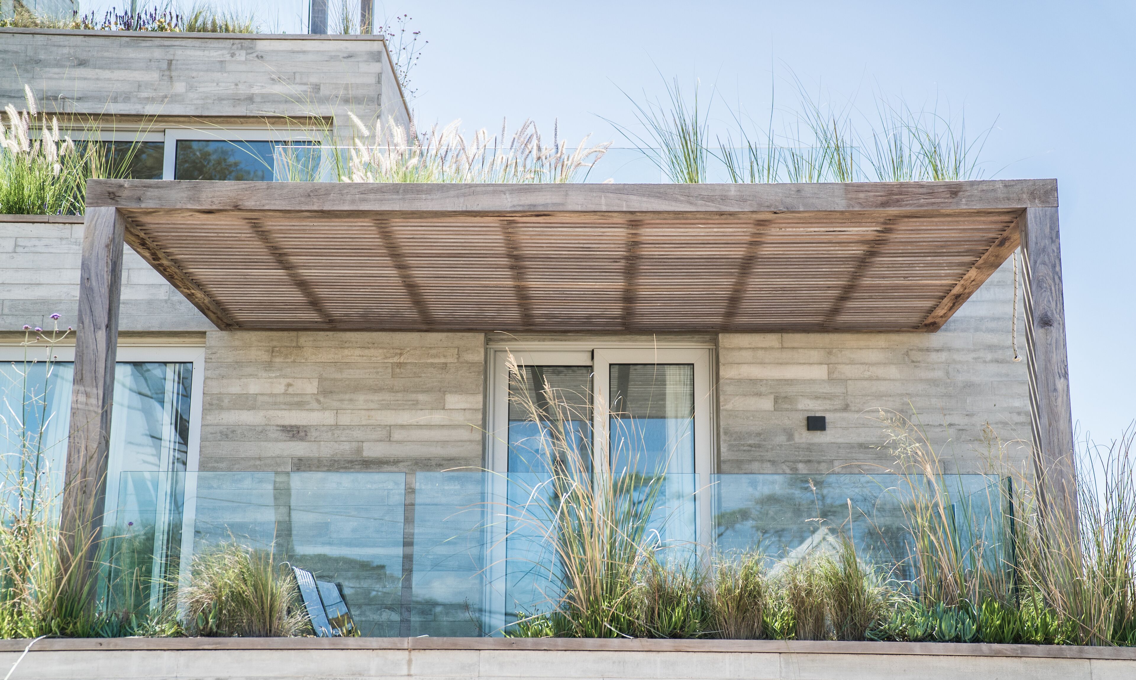 A modern building with a wooden pergola, large windows, and surrounding vegetation.