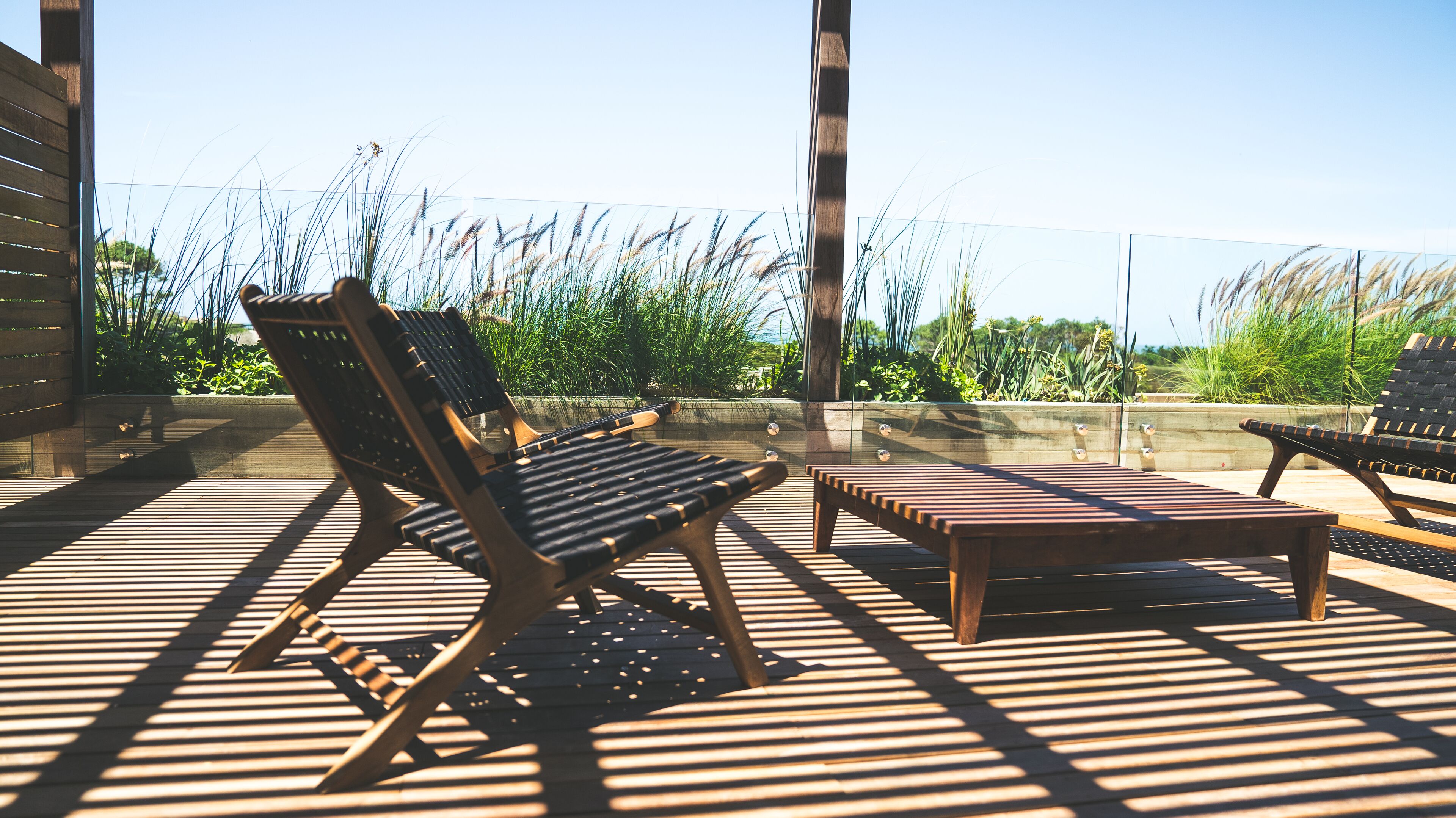 Eine Holzterrasse mit zwei Liegestühlen und einem kleinen Tisch, umgeben von hohem Gras und blauem Himmel.